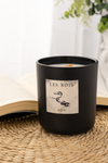 Black candle labeled 'Les Bois' on a woven surface with a blurred background